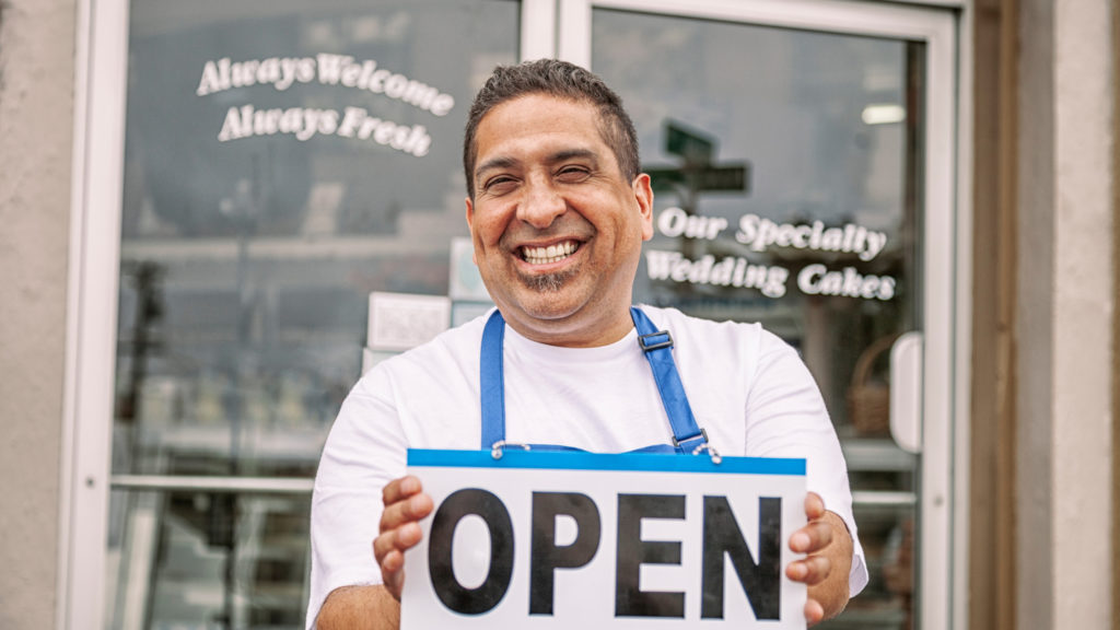 A hispanic man holding an open sign in front of a bakery